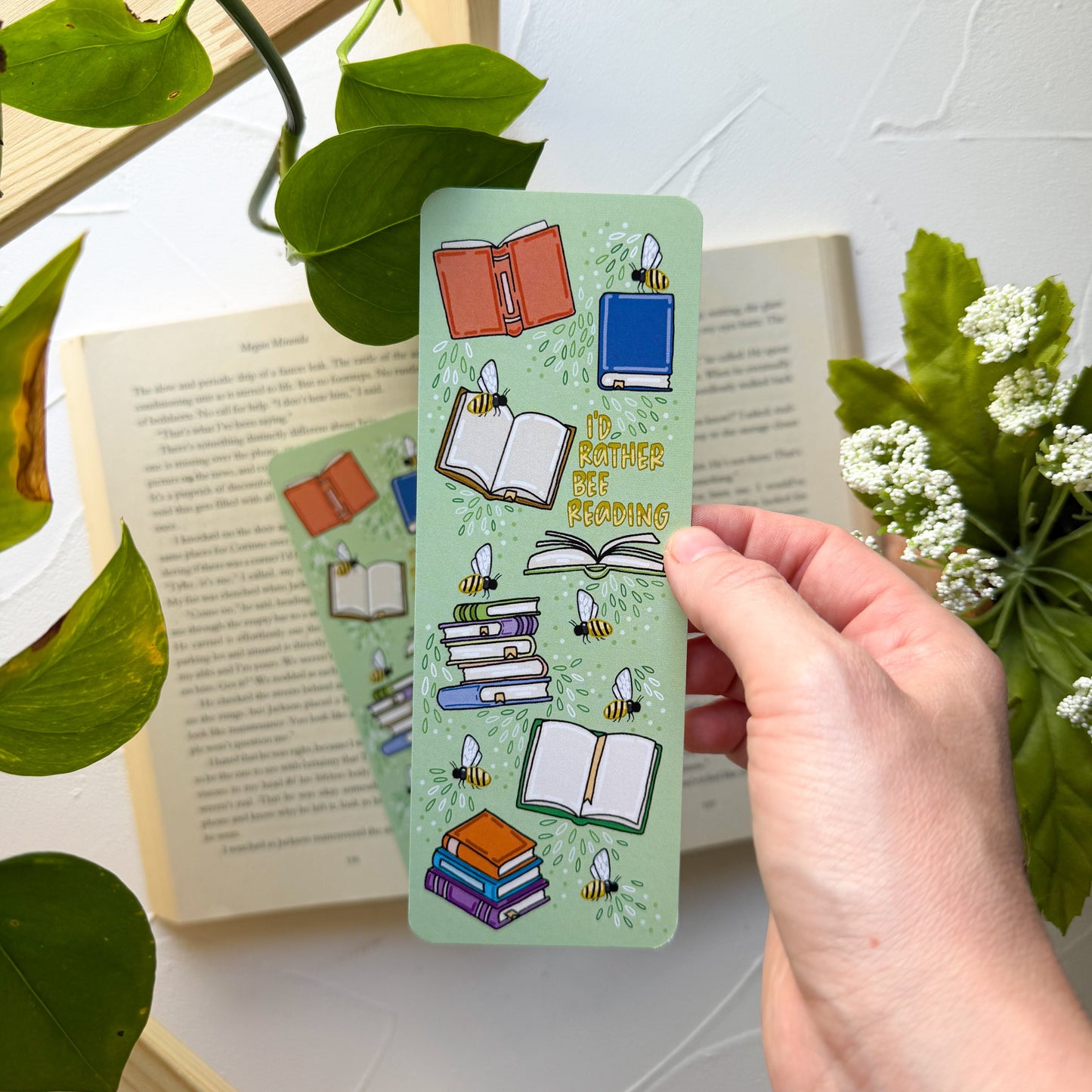 Hand holding a green bookmark with book illustrations and text, surrounded by books and plants.