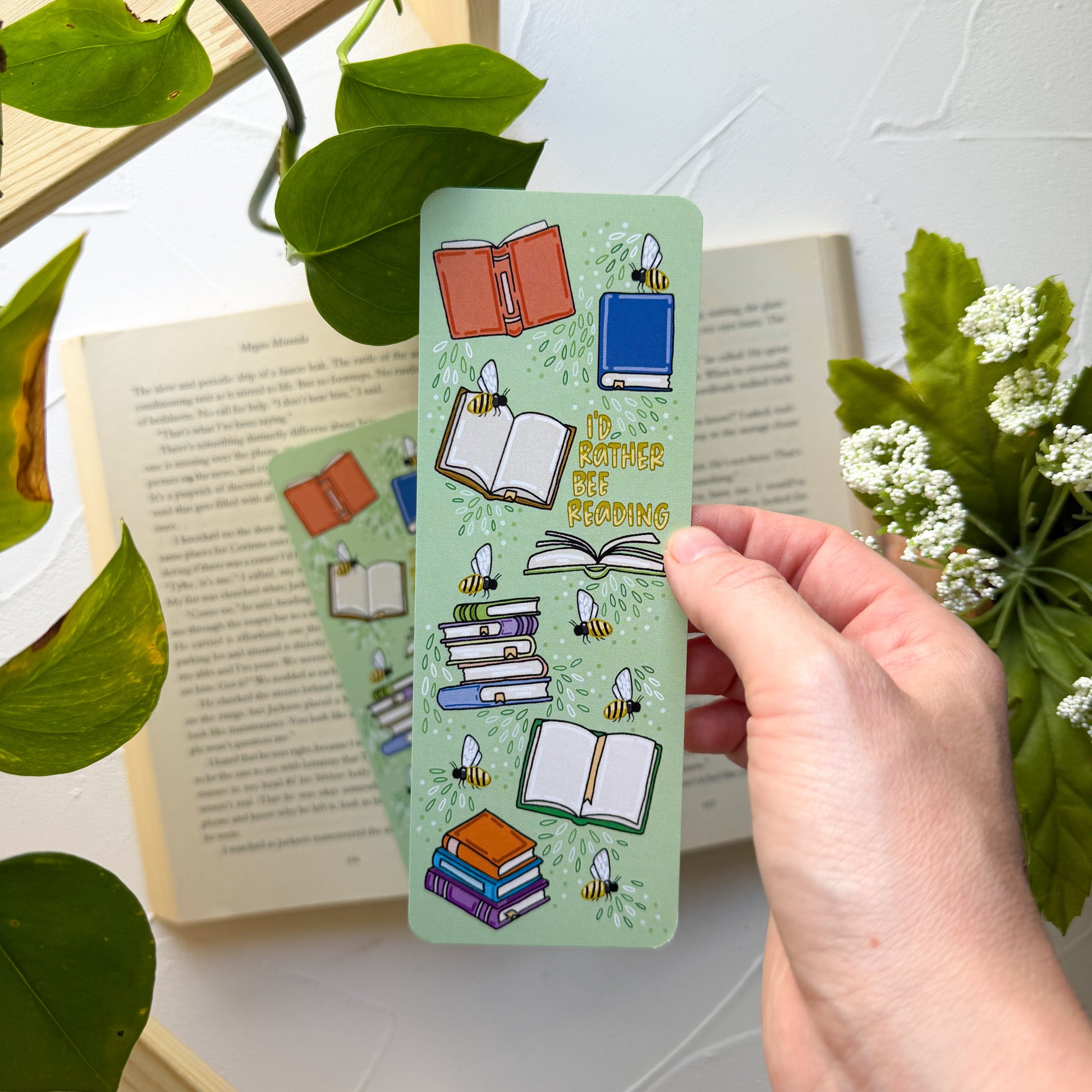 Hand holding a green bookmark with book illustrations and text, surrounded by books and plants.