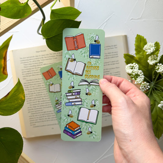 Hand holding a green bookmark with book illustrations and text, surrounded by books and plants.
