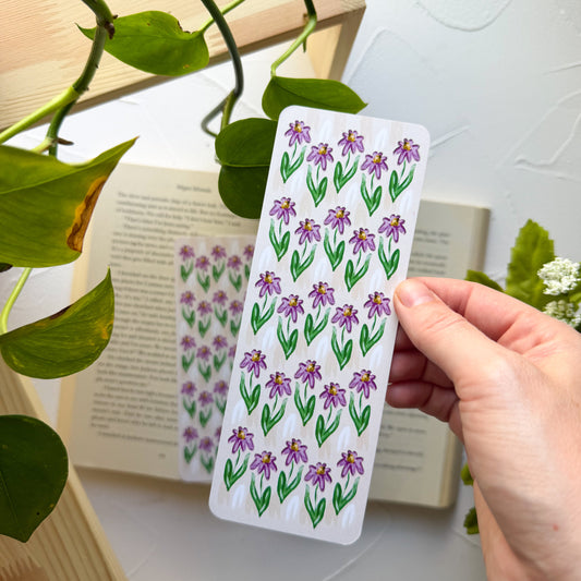 Hand holding a floral bookmark with a book and green leaves in the background