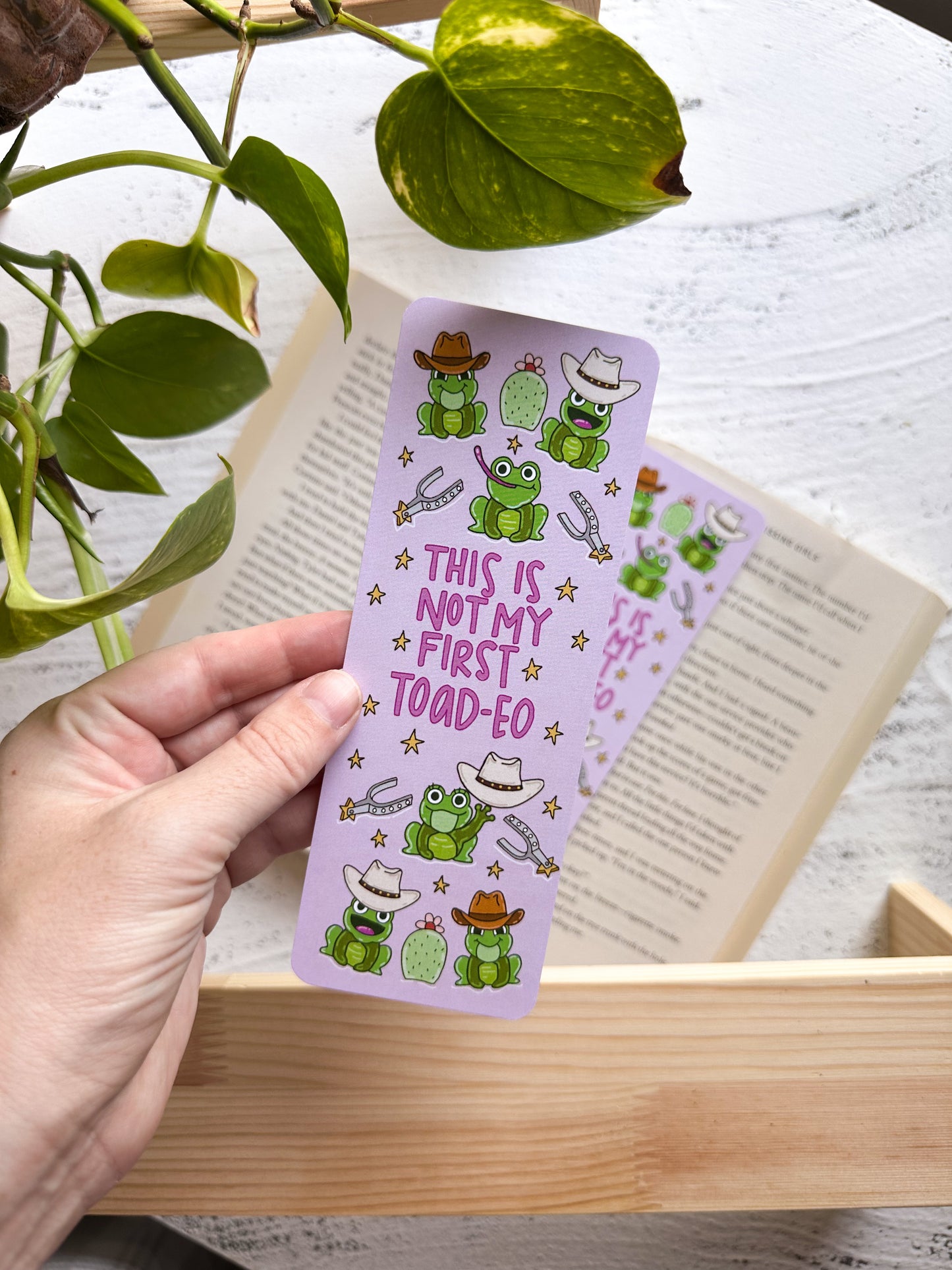 Hand holding a purple bookmark with frog illustrations and text, placed on an open book.