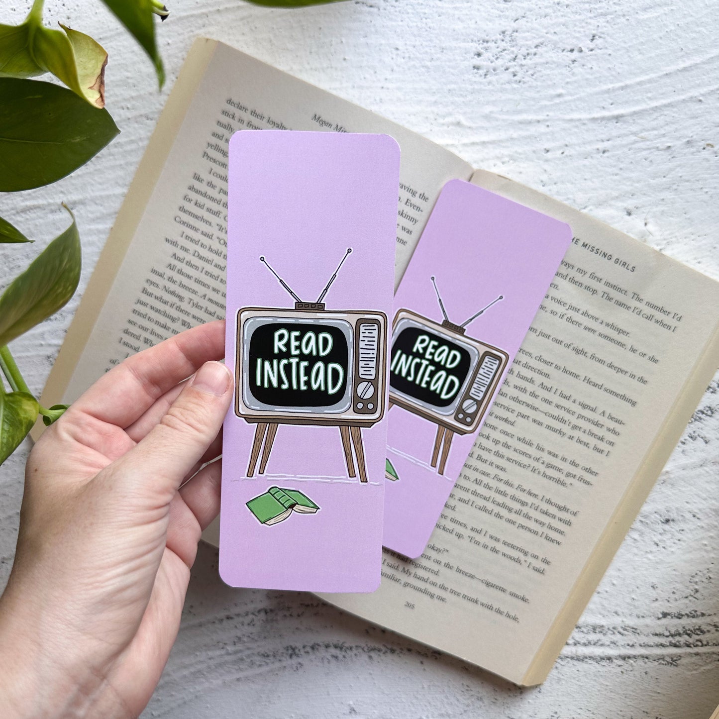 Pink bookmarks with vintage TV design and 'Read Instead' text held over an open book.