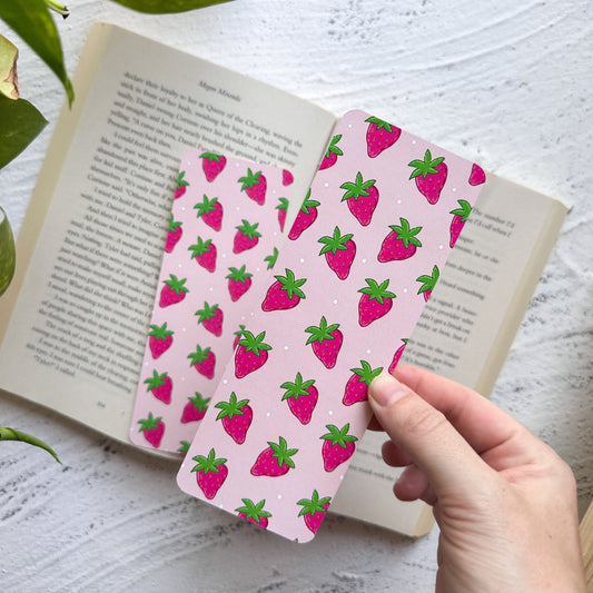 Two pink strawberry-patterned bookmarks on an open book with a hand holding one of them.