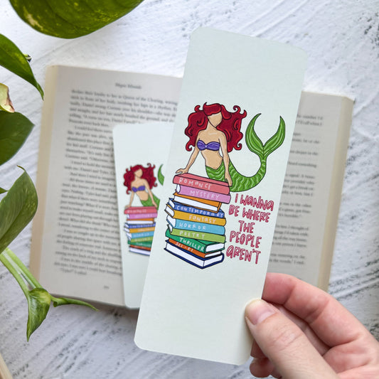 Hand holding a colorful mermaid bookmark with books on a white surface.